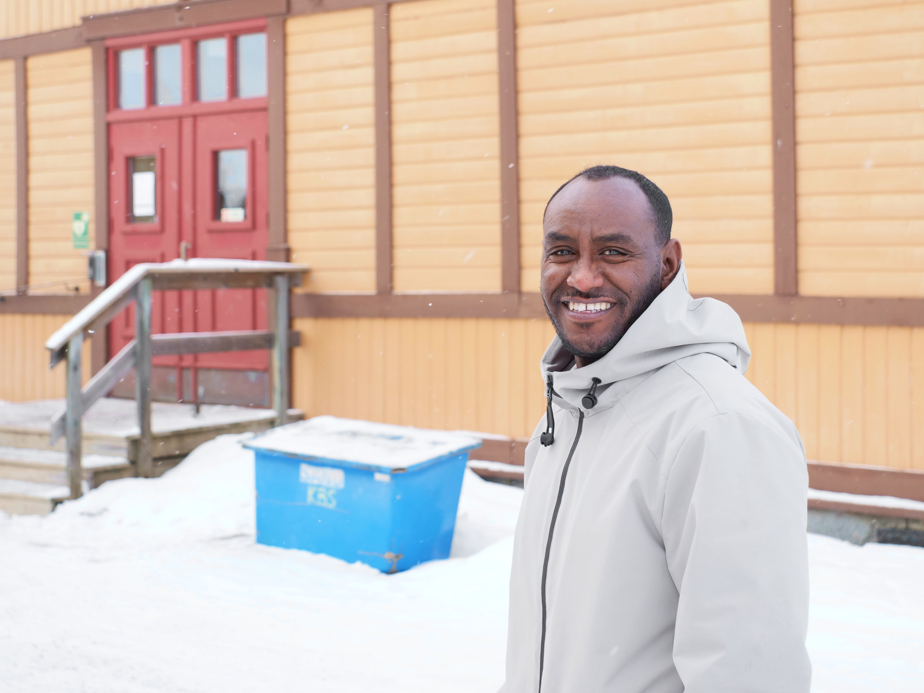 En leende man står framför gul byggnad i snöfall