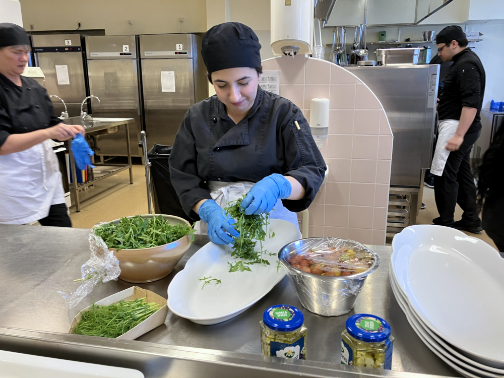 En person i kockkläder lägger upp sallad på ett fat i ett kök där det rör sig andra personer