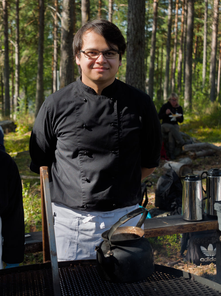 En person i kockkläder står vid en grillplats i en skog