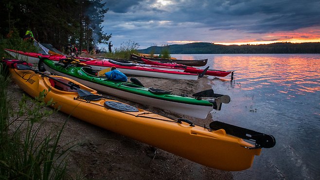 Bilden visar flera kanoter uppradade på en strand i Ångermanälven, solen håller på att gå ner 